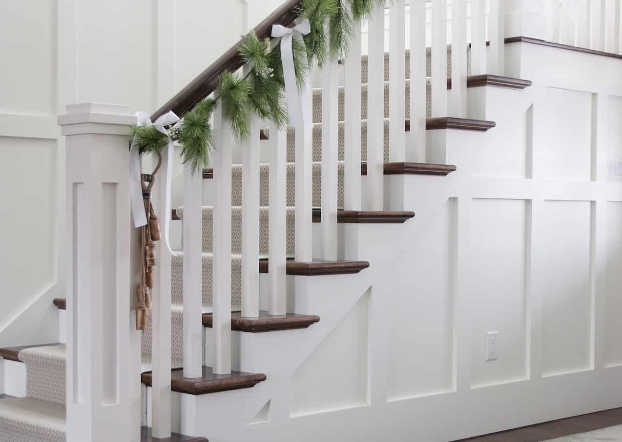 Neutral textured Christmas Home Tour 2019 from Caroline on Design. Stairs with white wainscoting, faux garland, sleigh bells, and white 2" ribbon.