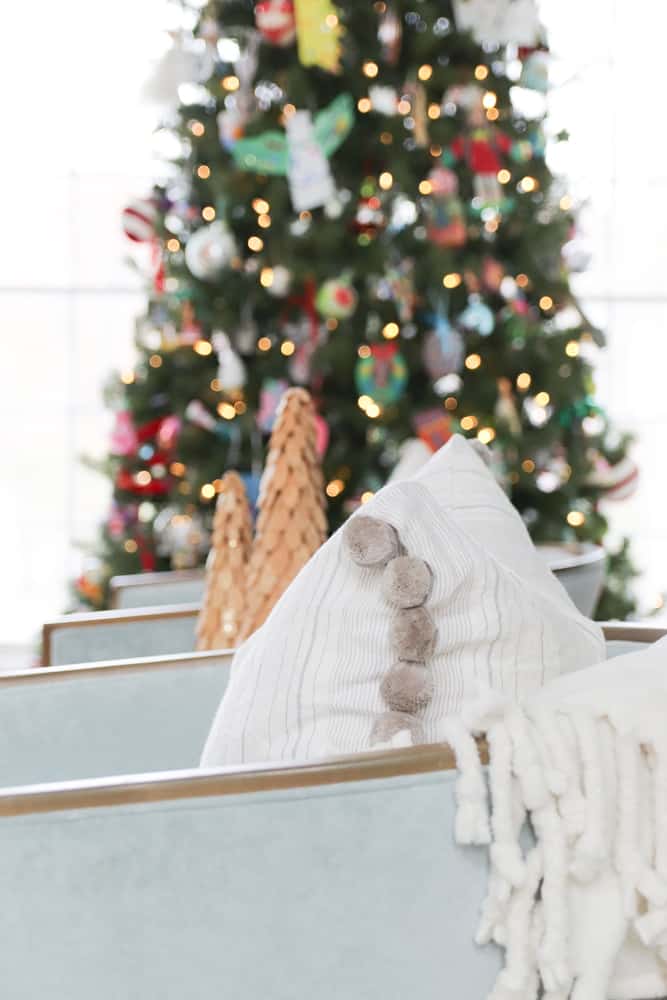Cozy pom throw pillow with soft white blanket on a blue velvet chair. Christmas tree lights twinkling in the background.