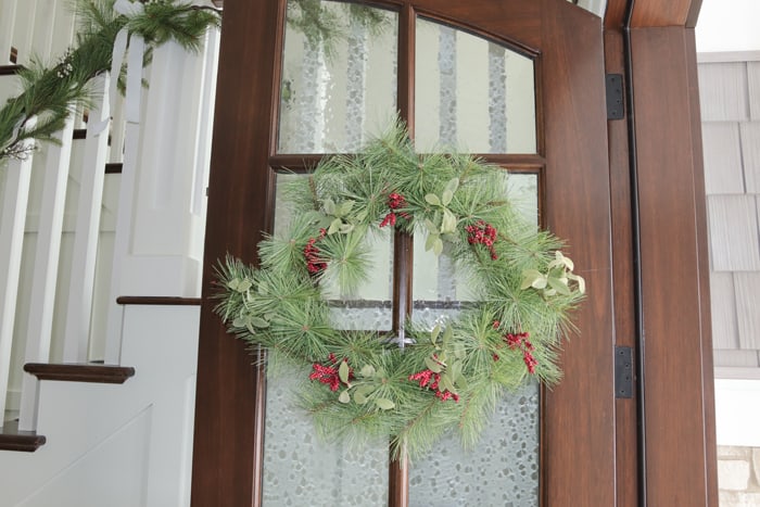 Christmas wreath with red berries hanging on real wood front door with seeded glass windows. Garland on stair railing in the background.