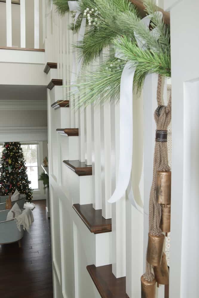 Faux garland tied with a white ribbon and sleigh bells on white and wood stairs.