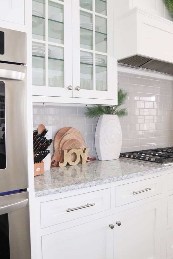 White kitchen with glass cabinet doors and grey backsplash tile. Simple neutral Christmas decorations on the grey quartz countertop.