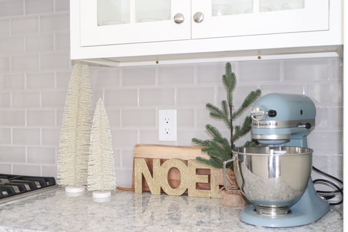 White kitchen cabinet with grey subway backsplash tile and neutral Christmas decor and blue Kitchenaid mixer.