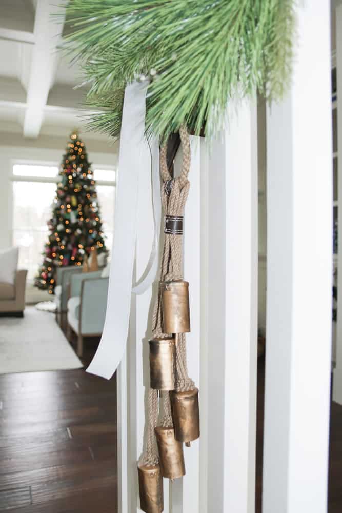 Close up of faux garland with white ribbon and sleigh bells on stairs. Twinkly Christmas tree with lights in the background.