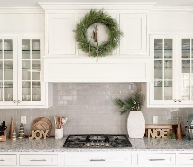 white kitchen with gray subway tile backsplash and faux wreath on stove hood with tan velvet ribbon and sleigh bells